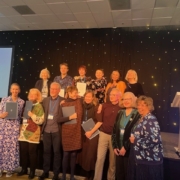 A group of people poses on stage at the SRHE International Research Conference. They hold documents, smiling under a spotlighted backdrop.
