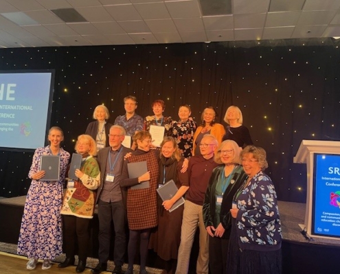 A group of people poses on stage at the SRHE International Research Conference. They hold documents, smiling under a spotlighted backdrop.