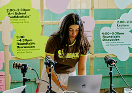 A person stands at a laptop, surrounded by microphones. Brightly colored signs in the background display event times and titles.