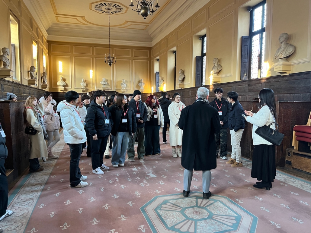 A group of people stand in a historical room with sculptures, ornate ceiling, and vintage décor, possibly in a museum or university.