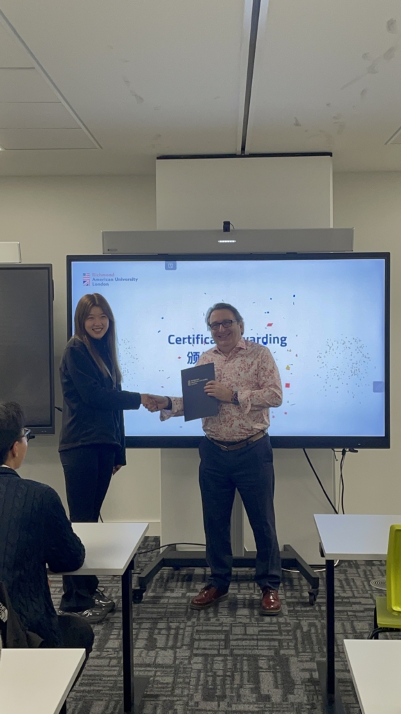 Two people exchange a certificate, shaking hands in a classroom. A screen displays "Certificate Awarding" with a logo in the background.