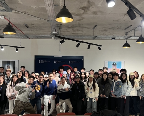 A group of people gather and pose indoors at Richmond American University London, with casual attire under modern ceiling lights.