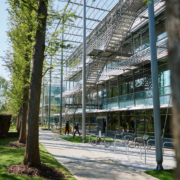 A modern building with glass windows stands beside a tree-lined path. People walk near bike racks on a sunny day.