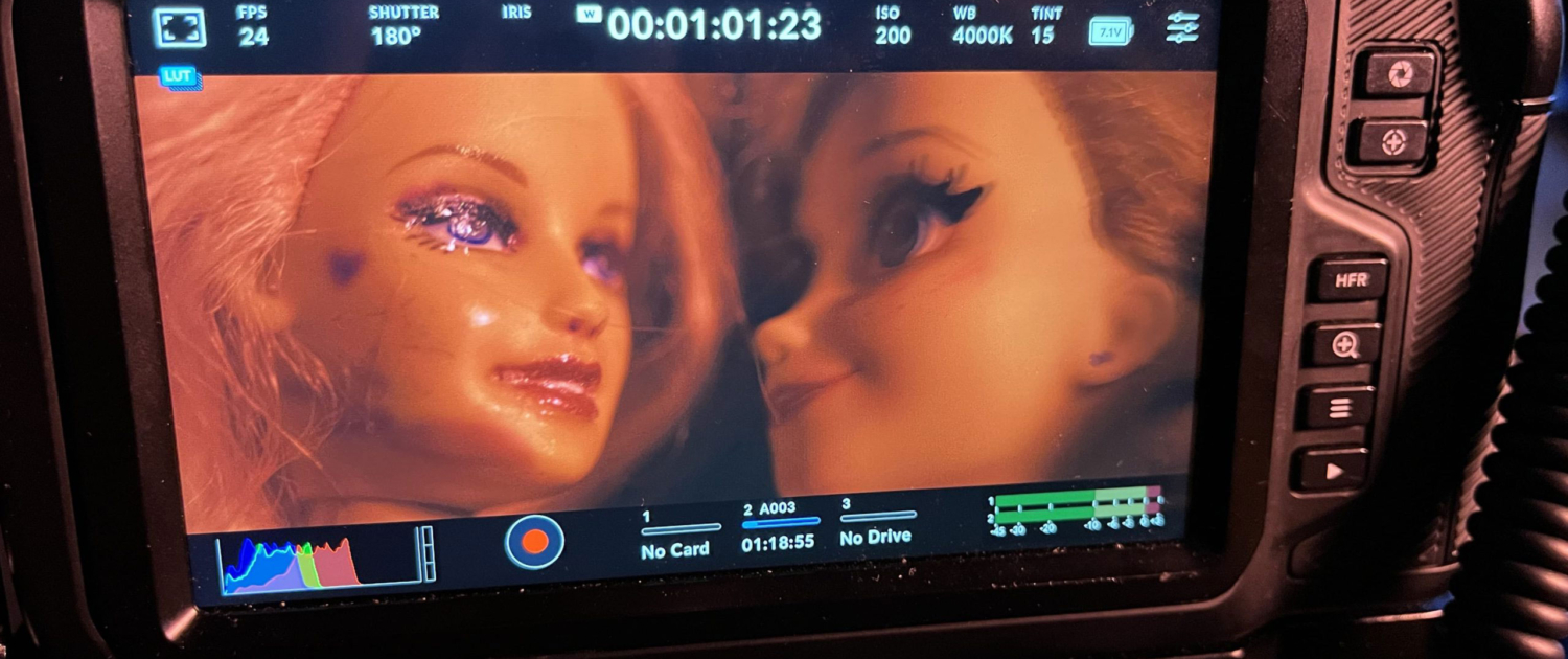 A camera screen displays a close-up of two doll faces with makeup, surrounded by the camera’s control interface and buttons.