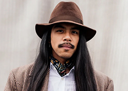 A person with long hair, wearing a brown hat, scarf, and checkered blazer, poses confidently against a neutral background.